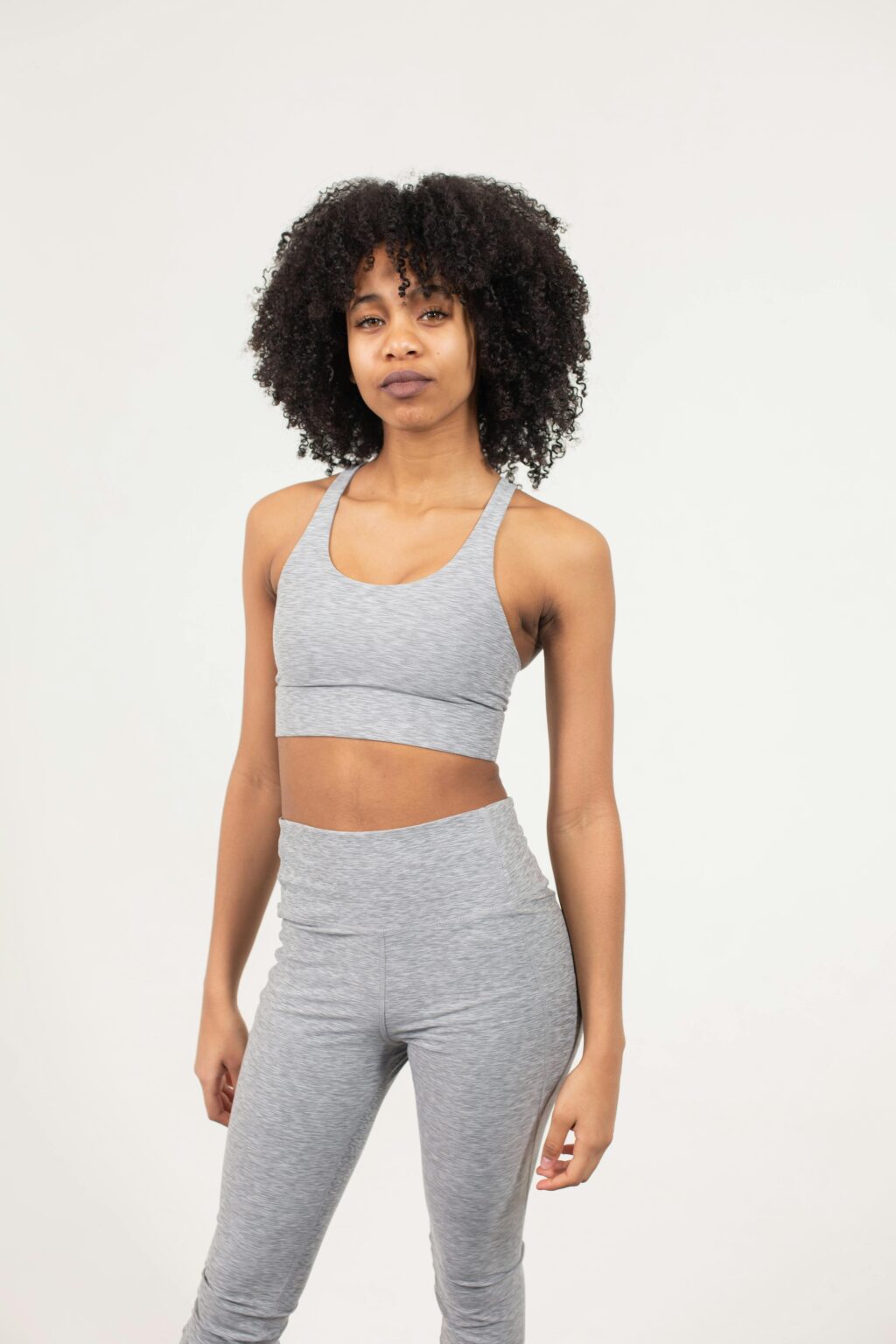 Emotionless slim African American female wearing gray sports bra and leggings standing on white background and looking at camera